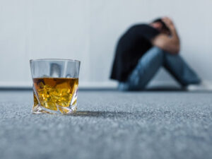 Glass of whiskey in focus with blurred man in background, depicting struggle with alcoholism.