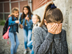 Upset girl covering her face as peers laugh in the background, implying bullying.