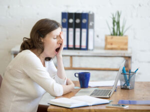 Woman yawning at her desk with a laptop, books, and a blue mug, showing fatigue.