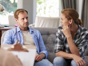 A man and woman sit closely on a couch, conversing in a cozy, sunlit room.