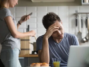 A man appears stressed in front of a laptop in a kitchen, with a woman in the background.