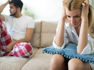 Woman upset on couch with man in background, suggesting conflict or concern.