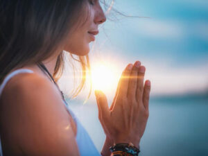 Woman in contemplative prayer with hands together against a sunset backdrop.