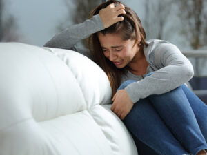 Distressed woman crying and clutching her head, seated on a couch.